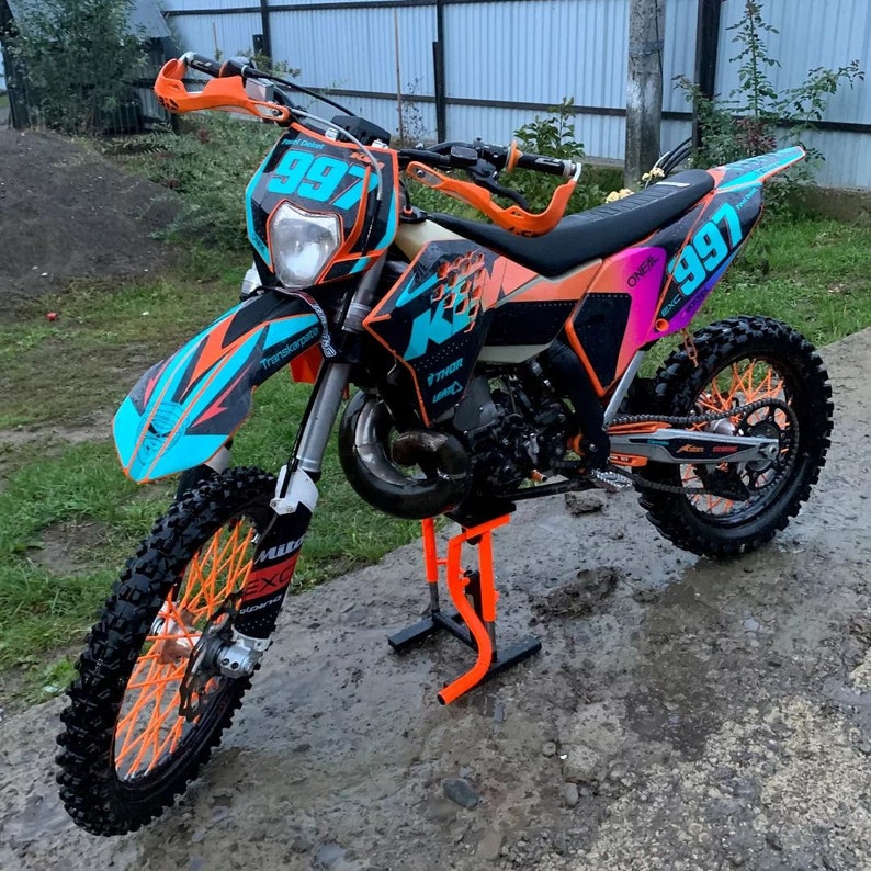 May include: A KTM dirt bike featuring a black, orange, and bright blue color scheme with the number 997 displayed on the front and side panels. The motorcycle is mounted on a bright orange metal stand and has orange wheel spokes and hand guards.