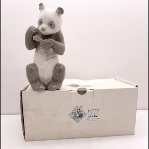 Puede incluir: Una figurita de porcelana blanca y gris de un panda sentado y sosteniendo un trozo de bambú en sus patas. La figurita está sobre una caja blanca con la palabra "NAO" impresa en ella.