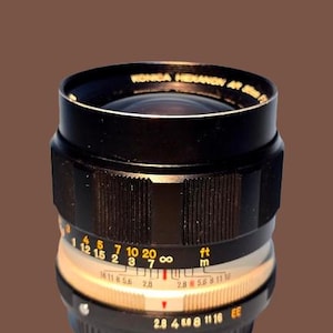 May include: A black Konica Hexanon AR 50mm F1.8 camera lens. The lens has a ribbed focus ring, distance markings in feet and meters, and aperture settings. The lens has a black exterior with gold and white markings.