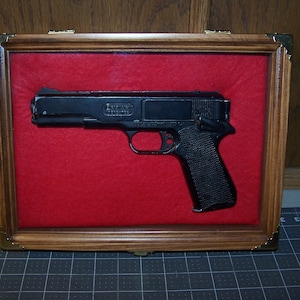 May include: A black air pistol displayed in a wooden shadow box with a red velvet lining. The pistol has a black plastic grip and a metal barrel.