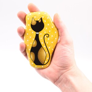 Op de afbeelding: Een hand houdt een geel geverfde steen vast met witte stippen. Op de steen is een zwarte kat met gouden vlekken geschilderd.