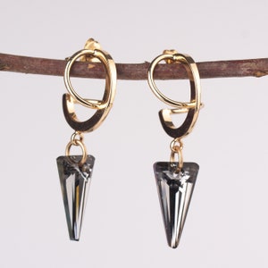 Op de afbeelding: Een paar gouden creolen met een hangende grijze kristallen traan. De oorbellen hangen aan een tak.