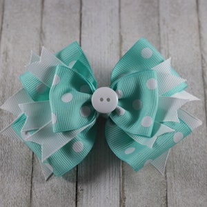 May include: A teal and white polka dot hair bow with a white button in the center. The bow is made of two layers of ribbon and is tied in a knot.