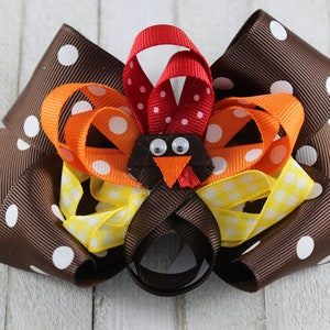 May include: A handmade hair bow in the shape of a turkey. The bow features layers of brown, orange, yellow, and red ribbons. The turkey's face has googly eyes and an orange beak. The bow is decorated with white polka dots and yellow gingham.