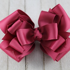 May include: A large, layered hair bow in a deep magenta color. The bow is made of grosgrain ribbon and has a textured appearance. The bow is centered and sits on a light-colored wooden surface. This accessory is suitable for various occasions.