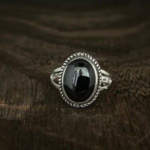 May include: A silver ring with a black oval gemstone set in a rope-like border.