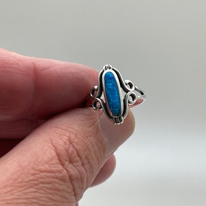 May include: Silver ring with an oval, blue opal center stone. The ring features decorative scrollwork on the sides of the opal and a simple band. The opal has a vibrant, blue color.