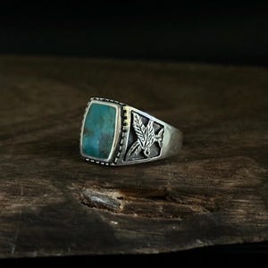 Op de afbeelding: Een zilveren ring met een rechthoekige turkooizen steen. De ring heeft een gedetailleerd ontwerp op de band, inclusief een bladmotief. De ring wordt gepresenteerd op een donkere houten ondergrond.