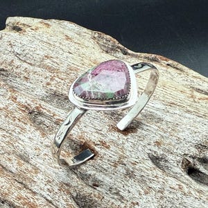 May include: A silver cuff bracelet with a heart-shaped gemstone in pink and green. The gemstone is set in a bezel setting. The bracelet has etched floral designs on the band.