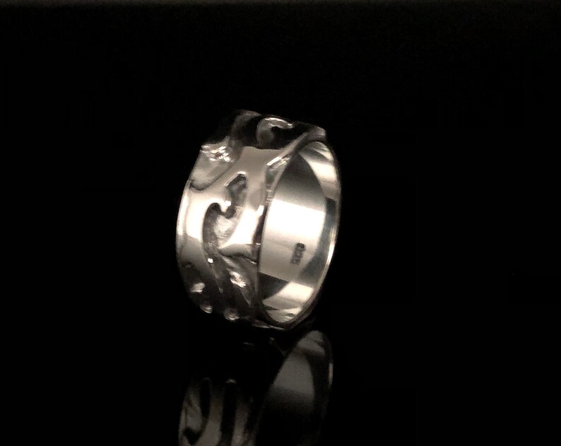 May include: A silver ring with a decorative, intricate design. The ring is wide and has a smooth, polished finish.