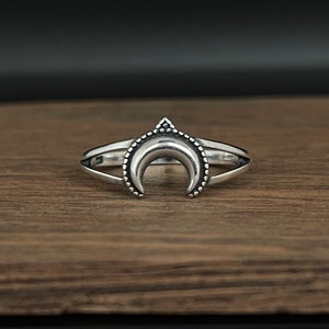 Peut inclure: Bague argent&eacute;e repr&eacute;sentant un croissant de lune. La bague a un double anneau et un petit triangle au sommet de la lune. La lune est orn&eacute;e de petites perles rondes le long de la courbe.