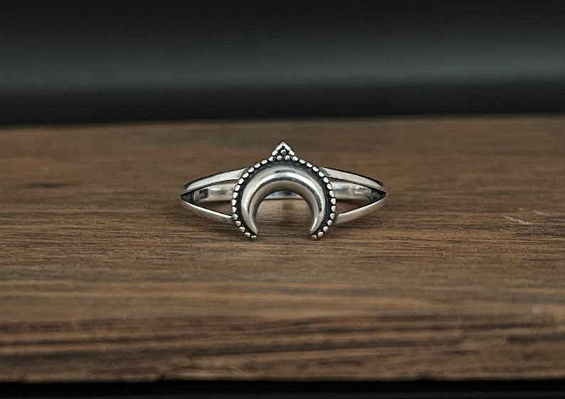 Peut inclure: Bague argent&eacute;e repr&eacute;sentant un croissant de lune. La bague a un double anneau et un petit triangle au sommet de la lune. La lune est orn&eacute;e de petites perles rondes le long de la courbe.