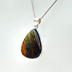 May include: A silver pendant with a teardrop-shaped gemstone. The stone displays a blend of colours: blue, gold, and brown. The pendant hangs from a silver chain with a twisted rope design. The gemstone is polished.