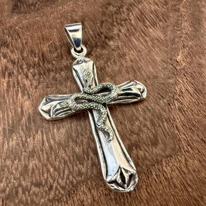 May include: A polished silver cross pendant featuring a textured, coiled snake wrapped around the centre. The cross has a smooth, reflective finish with flared ends and a sturdy bail at the top for attachment to a chain or cord necklace.