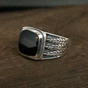 Könnte beinhalten: Ein silberner Ring mit einem schwarzen Stein und einem detaillierten Federdesign auf der Band.