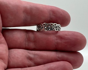 May include: A silver-colored ring with a Celtic knot design. The ring features an intricate pattern of interwoven lines, creating a continuous, symmetrical design. The ring is held between fingers, showcasing its detailed craftsmanship.