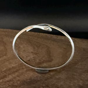May include: A silver-colored bangle bracelet with a snake head design. The bracelet is circular, with the snake's head at the top, and is resting on a wooden surface. The snake's head has small, detailed eyes.