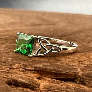 May include: A silver-toned ring featuring a square-cut, vibrant green gemstone set in a prong setting. The band displays an intricate, open-work knot design on both sides of the central stone, creating a continuous metal pattern along the shank.