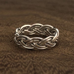 May include: A silver-colored ring with a braided design. The ring features a continuous pattern of interwoven strands, creating a textured and detailed appearance. The ring is shown on a brown surface.