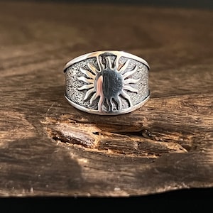 May include: A silver ring with a sun design. The ring features a textured band and a detailed sunburst in the center. The ring is displayed on a wooden surface.