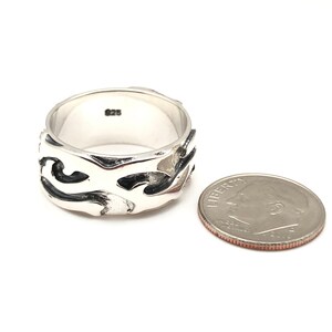 May include: A silver ring with a wide band featuring a black and silver abstract design. The ring is next to a US dime for size comparison.