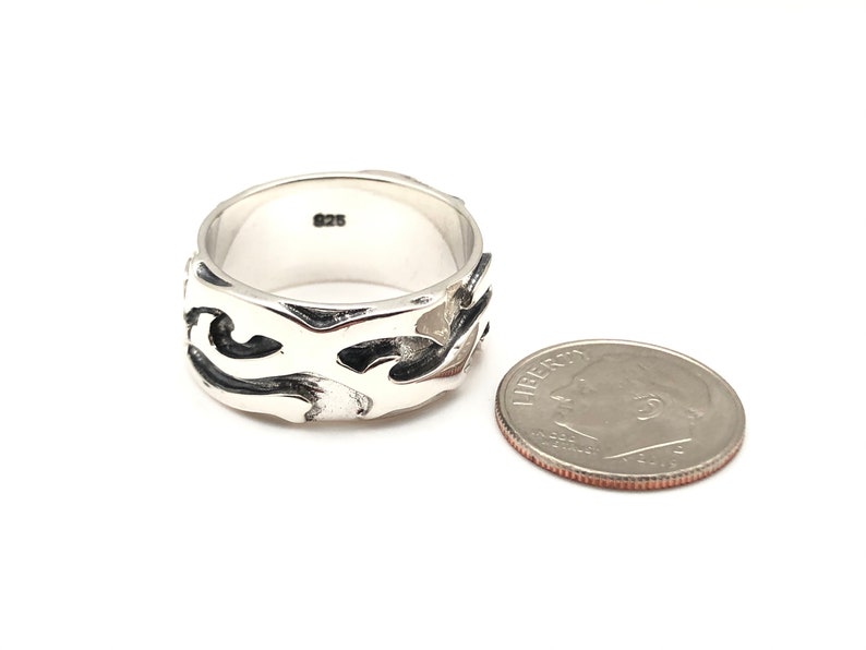 May include: A silver ring with a wide band featuring a black and silver abstract design. The ring is next to a US dime for size comparison.