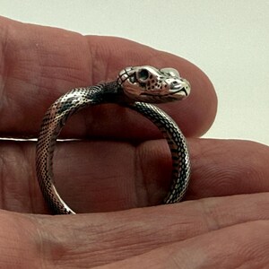 May include: A silver-toned ring shaped like a coiled snake with detailed scales and a defined head. The metal has a polished finish with darkened recessed areas to highlight the texture of the snake's skin and facial features, including eyes and mouth.