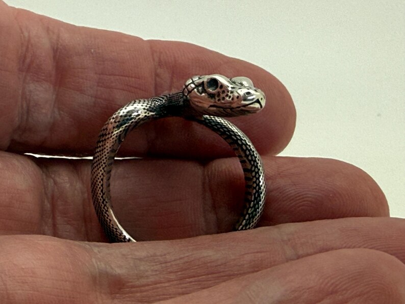 May include: A silver-toned ring shaped like a coiled snake with detailed scales and a defined head. The metal has a polished finish with darkened recessed areas to highlight the texture of the snake's skin and facial features, including eyes and mouth.