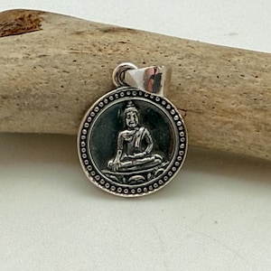 Puede incluir: Un colgante circular de plata que presenta un relieve de una figura de Buda sentado en el centro. El colgante tiene un fondo oscuro oxidado dentro de un borde de cuentas e incluye una anilla de plata pulida en la parte superior para la cadena.