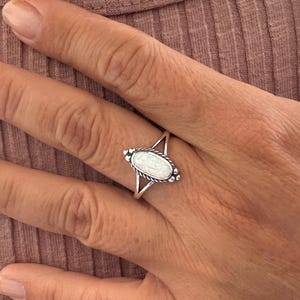 May include: A silver ring with an oval, white gemstone. The ring has a decorative design around the gemstone and a split band. The ring is on a hand with a pinkish-brown background.