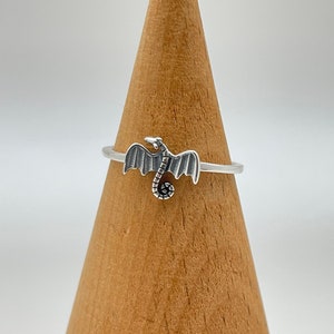 May include: A silver ring featuring a dragon design. The dragon has outstretched wings and a curled tail. The ring is displayed on a wooden cone against a white background. The ring is a delicate piece of jewelry.