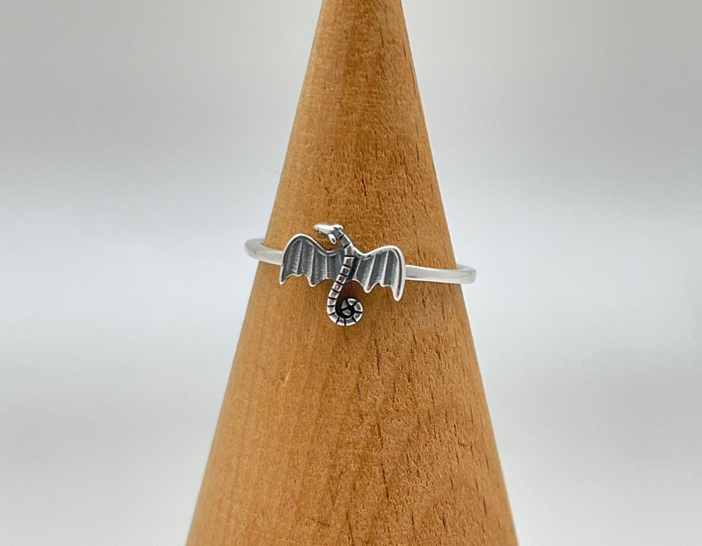 May include: A silver ring featuring a dragon design. The dragon has outstretched wings and a curled tail. The ring is displayed on a wooden cone against a white background. The ring is a delicate piece of jewelry.