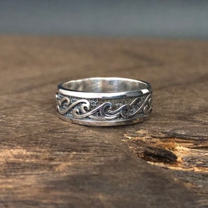 May include: A silver ring featuring a wave design. The ring is displayed on a wooden surface.