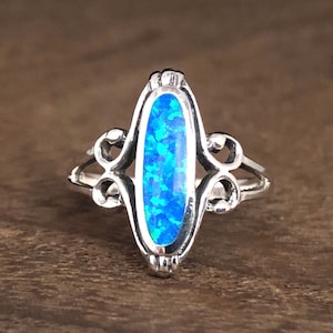 May include: A silver ring with an oval, blue opal stone. The opal has a mosaic pattern of light and dark blue. The ring has a decorative, openwork design on the sides and a detailed setting around the stone.