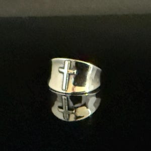 May include: A silver-colored ring with a cross design. The ring is wide and has a polished finish. The cross is centered on the front of the ring. The ring is reflected on the surface below.