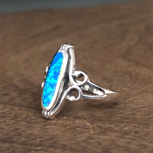May include: A silver ring with an elongated, oval-shaped blue opal stone. The ring features decorative filigree on the sides and around the stone, creating an elegant and detailed design. The opal has a vibrant, multi-toned blue color.