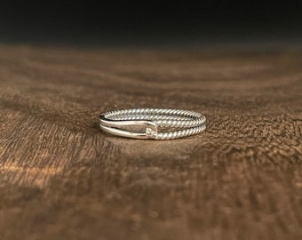 May include: A silver ring with a twisted rope design. The ring features two bands connected by a small, curved bar. The ring is set against a dark brown wooden surface.