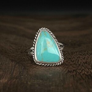 May include: A turquoise ring with a silver setting. The turquoise is a triangular shape and has a light blue color. The ring is set in a silver band with a rope-like design.
