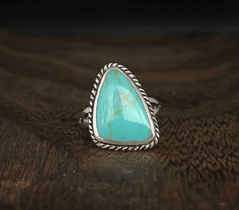 May include: A turquoise ring with a silver setting. The turquoise is a triangular shape and has a light blue color. The ring is set in a silver band with a rope-like design.