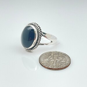 May include: A silver ring featuring a black oval gemstone set within a rope-like border. The ring is displayed alongside a UK ten pence coin for scale.