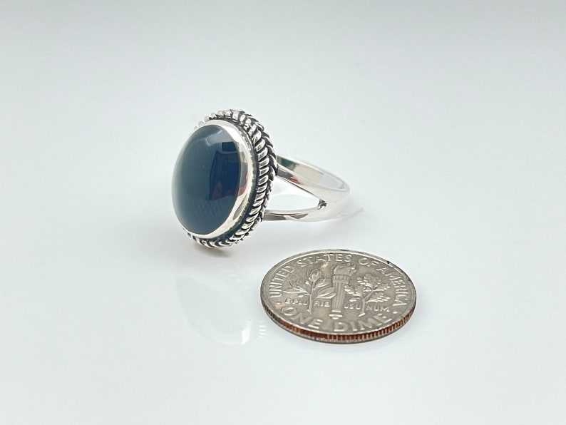 May include: A silver ring featuring a black oval gemstone set within a rope-like border. The ring is displayed alongside a UK ten pence coin for scale.