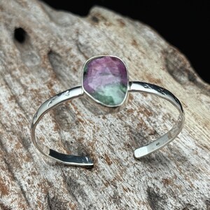 May include: A silver cuff bracelet with a large, oval gemstone. The gemstone has pink and green colours. The bracelet has an open design with etched detailing on the band. The bracelet is displayed on a piece of driftwood.