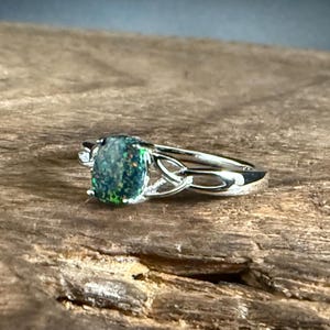 May include: A silver-toned ring featuring an oval-shaped dark green opal gemstone with iridescent flecks of red and blue. The metal band displays a decorative openwork design on both sides of the central stone, creating a continuous metallic loop.
