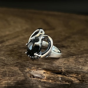 May include: A silver ring with a black gemstone set in a unique, intricate design. The ring features a swirling, organic shape that resembles a stylized heart.