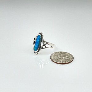 May include: A silver ring with an oval, blue opal center stone. The ring has a decorative design on the sides and a split shank. A dime is next to the ring for size comparison.