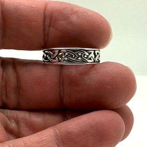 May include: A silver ring with a Celtic knot design. The ring features an intricate pattern of interwoven lines, creating a continuous, symmetrical design. The ring is approximately 0.25 inches wide.