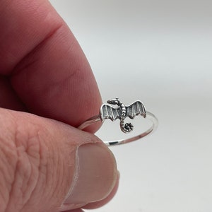 May include: A silver dragon ring. The ring features a detailed dragon with outstretched wings and a curled tail. The dragon is the focal point of the ring, with a polished finish. The ring is a delicate piece of jewelry.