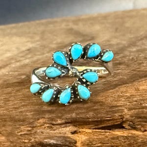 May include: A silver ring featuring a bypass design adorned with ten teardrop-shaped turquoise stones. The stones are set in individual bezels arranged in two curved rows that overlap at the center, creating a continuous openwork pattern.