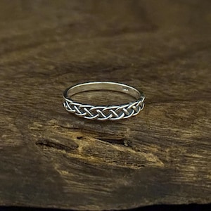 Puede incluir: Un anillo de plata con un diseño de nudo celta. El anillo presenta un patrón entrelazado alrededor de la banda. El anillo se muestra sobre una superficie de madera oscura.