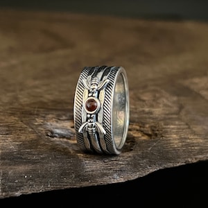 May include: A silver ring with a red gemstone set in the center. The ring has a textured design with a repeating pattern of lines and a small, stylized insect on each side of the stone.